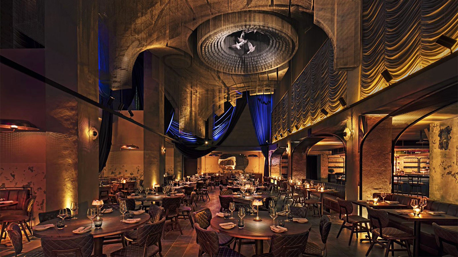 The dining room at Cathédrale with hanging sculptures evoking Gothic architectural elements, arches throughout the space, leather chairs and round wooden tables